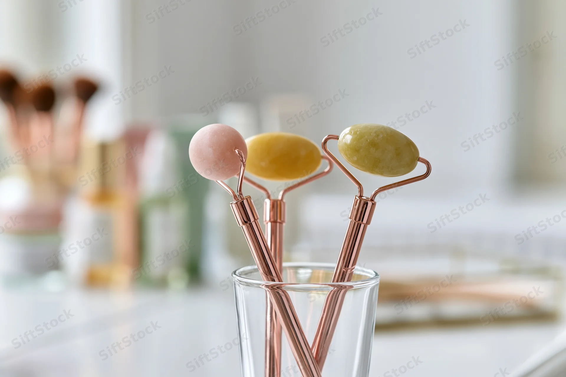 Three jade face rollers with rose gold handles standing in a glass cup on a vanity countertop