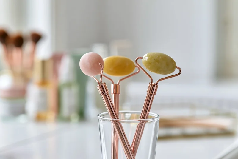 Three jade face rollers with rose gold handles standing in a glass cup on a vanity countertop