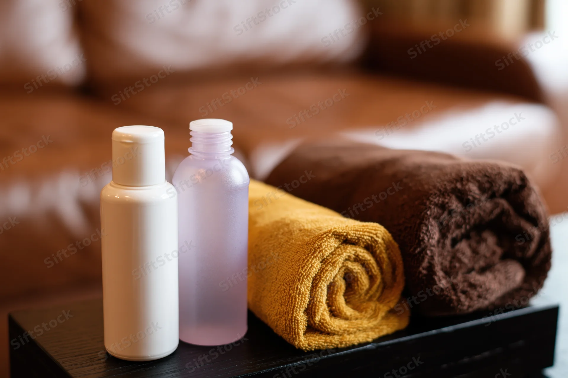 Two small hotel toiletry bottles and rolled towels on a tray beside a bed