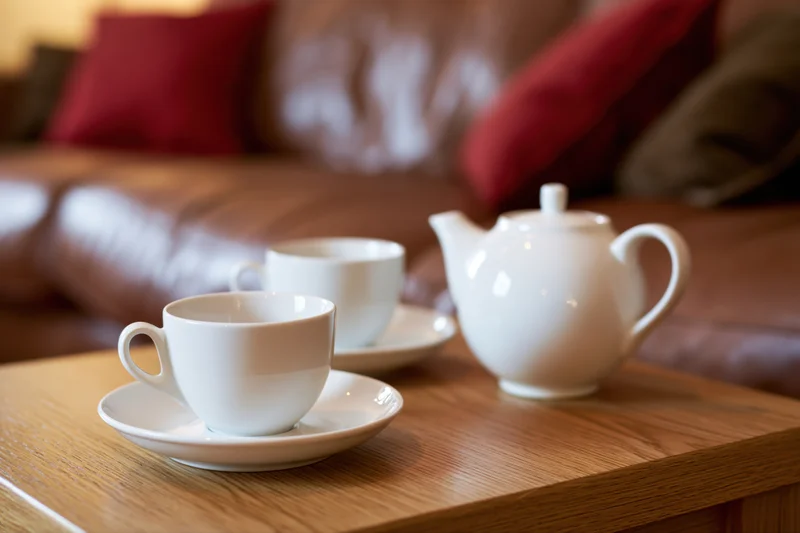 Two white ceramic teacups on saucers with a matching teapot on a wooden table in a living room with a brown sofa and red cushions in the background