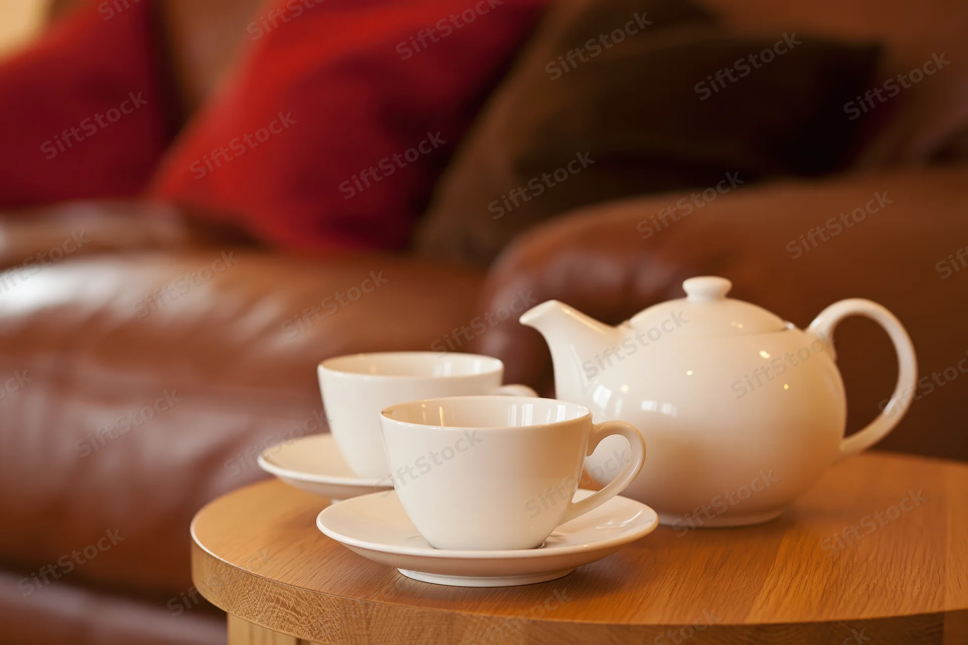 Two white ceramic teacups on saucers with a matching teapot on a wooden side table in a living room with a leather sofa and red cushions