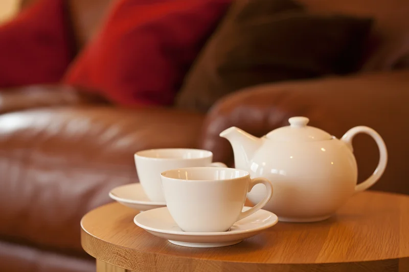 Two white ceramic teacups on saucers with a matching teapot on a wooden side table in a living room with a leather sofa and red cushions