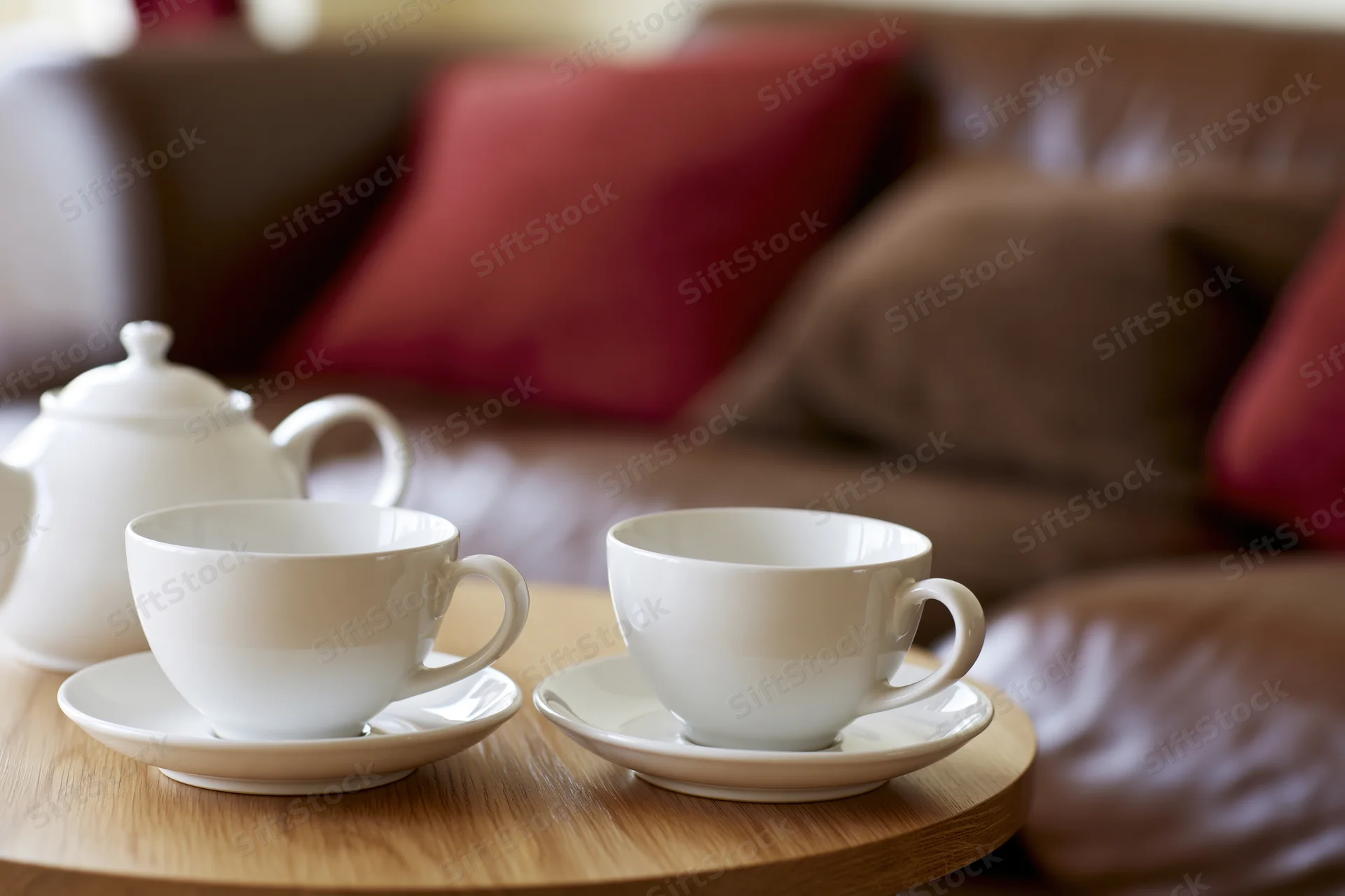 Two white ceramic teacups on saucers with a matching teapot on a wooden table in a living room with a brown sofa and red cushions in the background