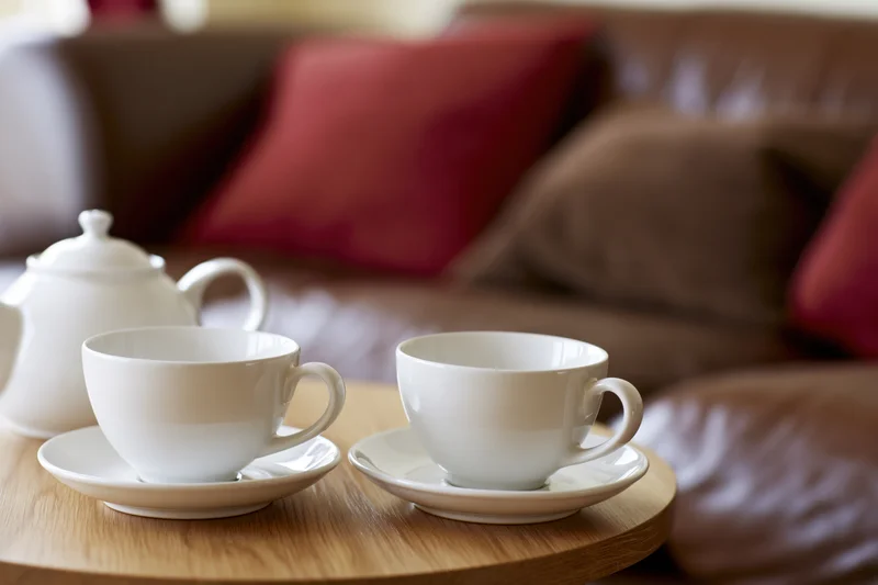 Two white ceramic teacups on saucers with a matching teapot on a wooden table in a living room with a brown sofa and red cushions in the background
