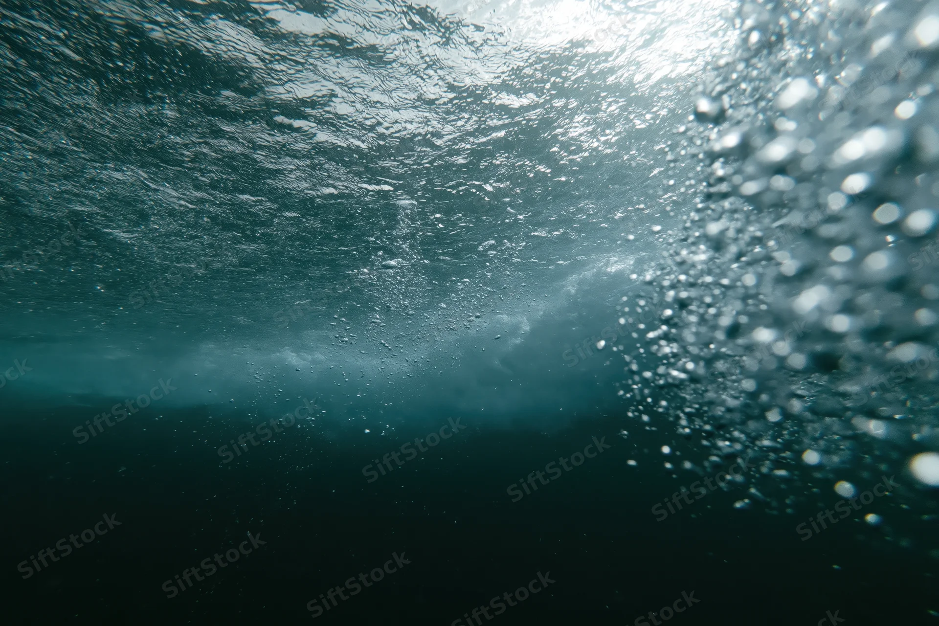 Underwater view looking up through the churning underside of a wave with sunlight filtering through the surface and rising bubbles