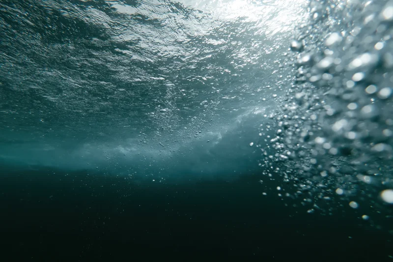 Underwater view looking up through the churning underside of a wave with sunlight filtering through the surface and rising bubbles