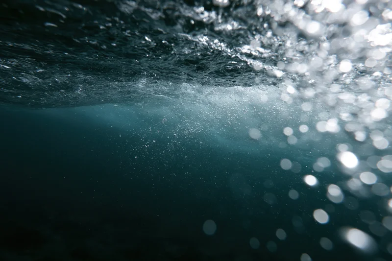 Underwater view looking up through the churning underside of a wave with sunlight filtering through bubbles and ripples