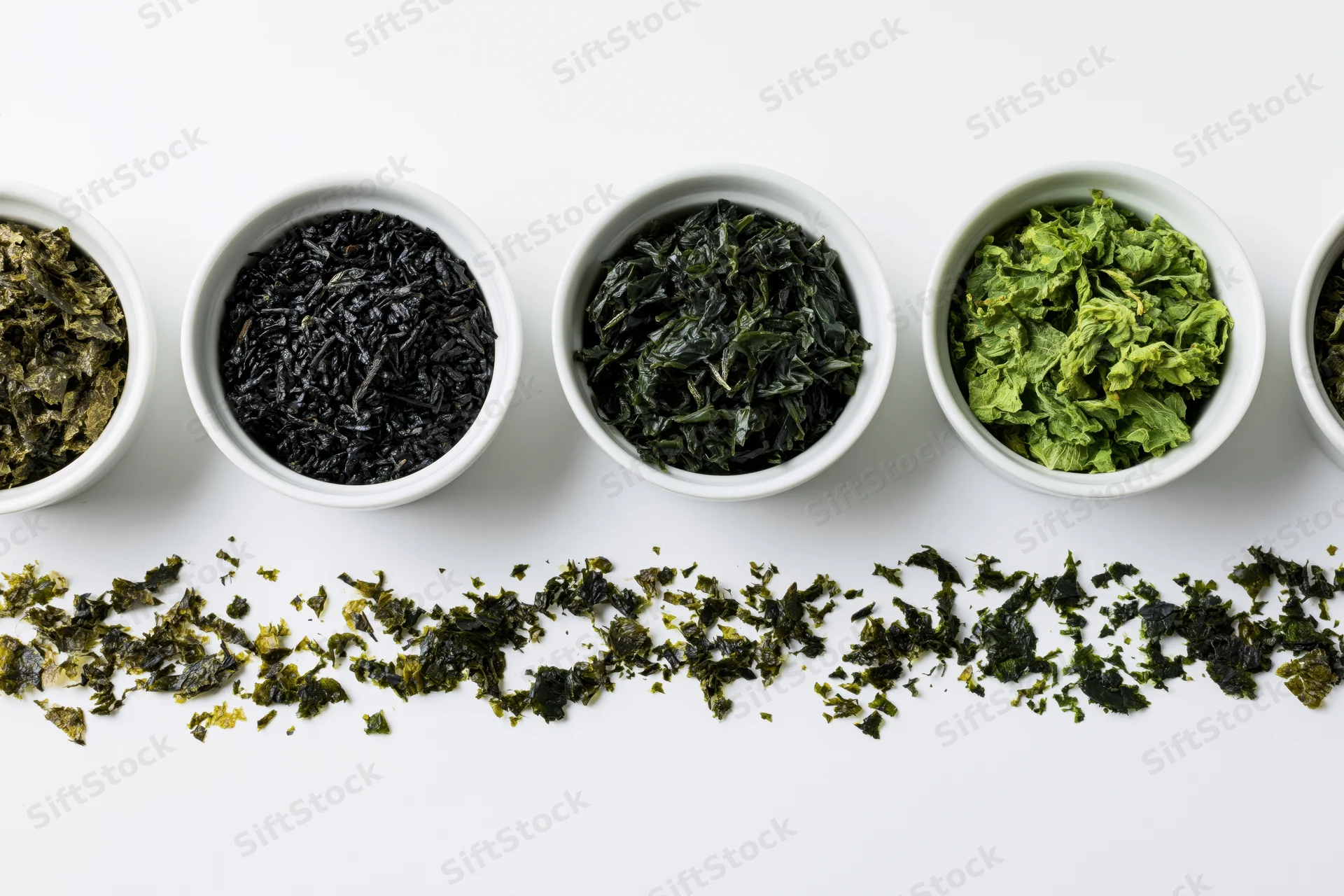 Various dried seaweed varieties in small white ceramic bowls arranged in a row on a white surface with scattered pieces below