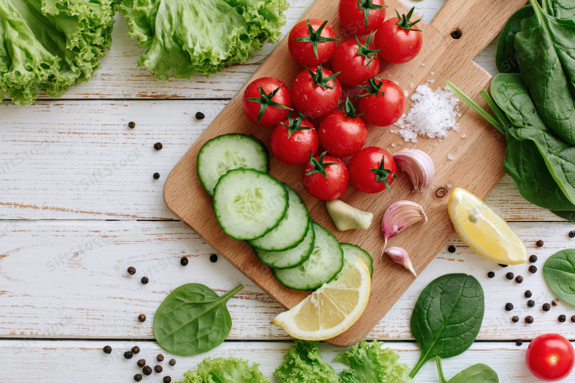 Cherry tomatoes, sliced cucumber, lemon, garlic, and spinach arranged on a wooden cutting board with lettuce and peppercorns on a white wooden surface