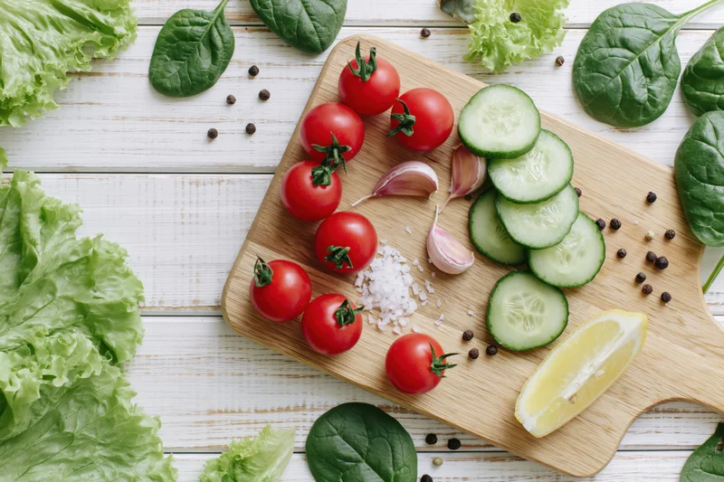 Cherry tomatoes, sliced cucumber, garlic, lemon, salt, pepper, lettuce, and spinach arranged on a wooden cutting board on a white wooden surface