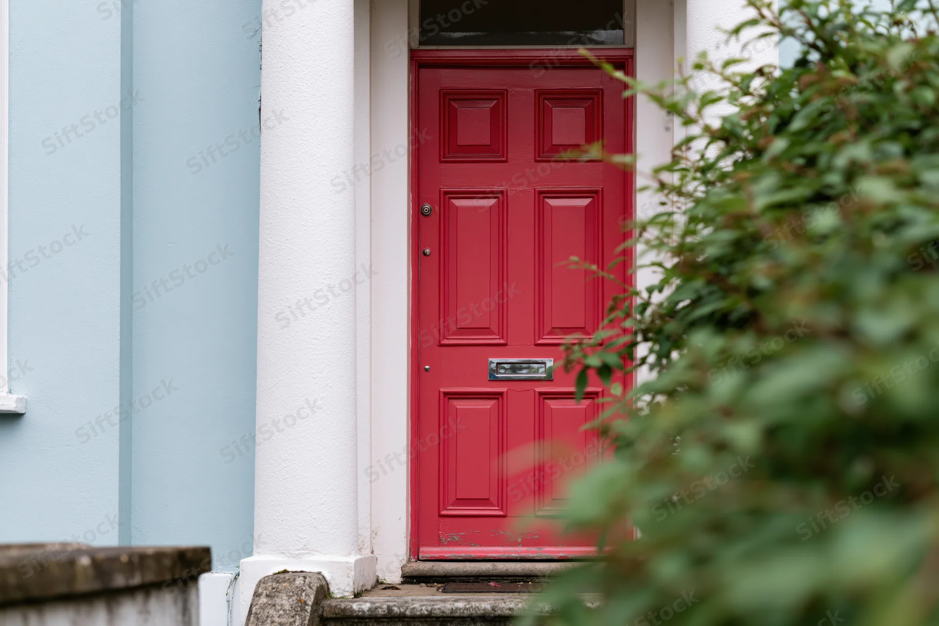 Bold red painted front door with raised panels and chrome mailslot on a house exterior framed by white columns and green foliage