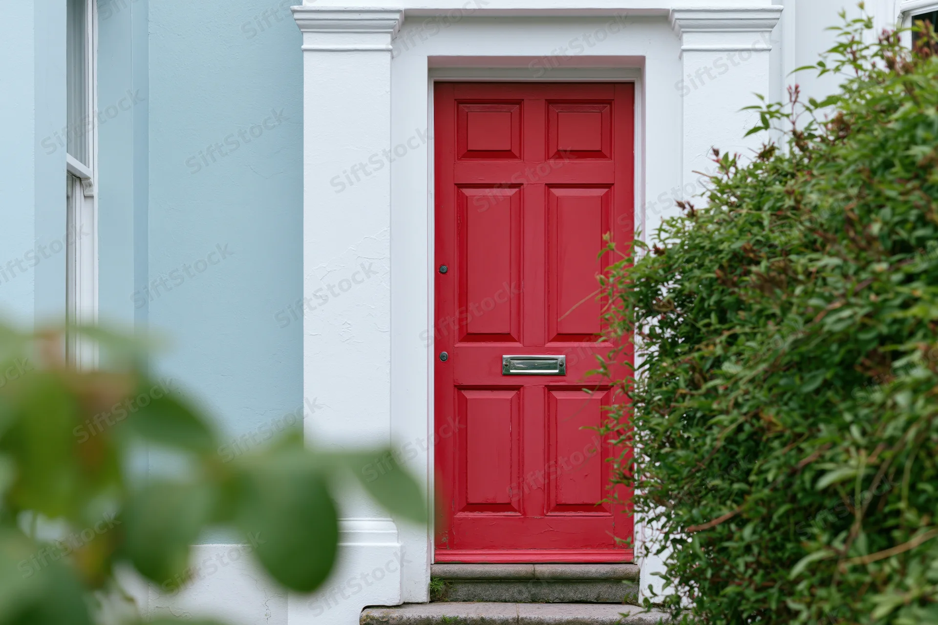 Bold red front door with raised panels and chrome mailslot on a white-framed house entrance surrounded by green foliage
