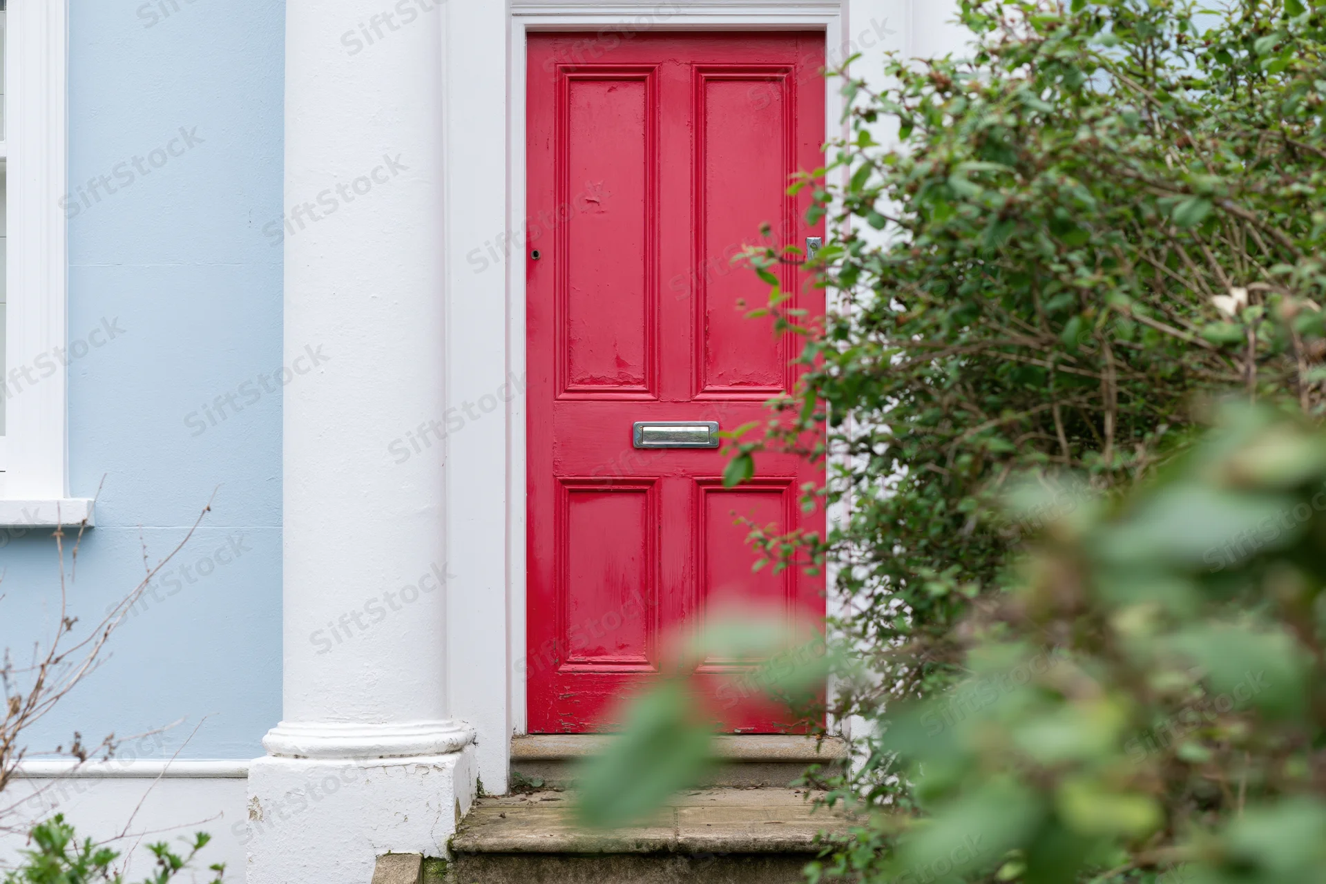 Red painted front door with raised panels and chrome mailslot on a blue house exterior framed by green foliage