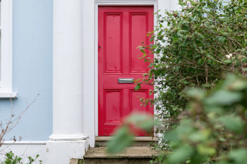 Red painted front door with raised panels and chrome mailslot on a blue house exterior framed by green foliage
