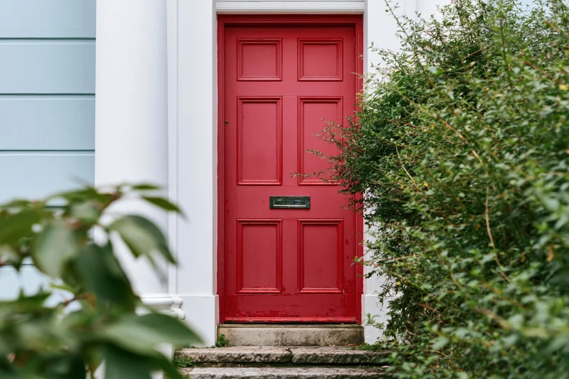 Red painted front door with raised panels and chrome mailslot on a white house exterior framed by green foliage