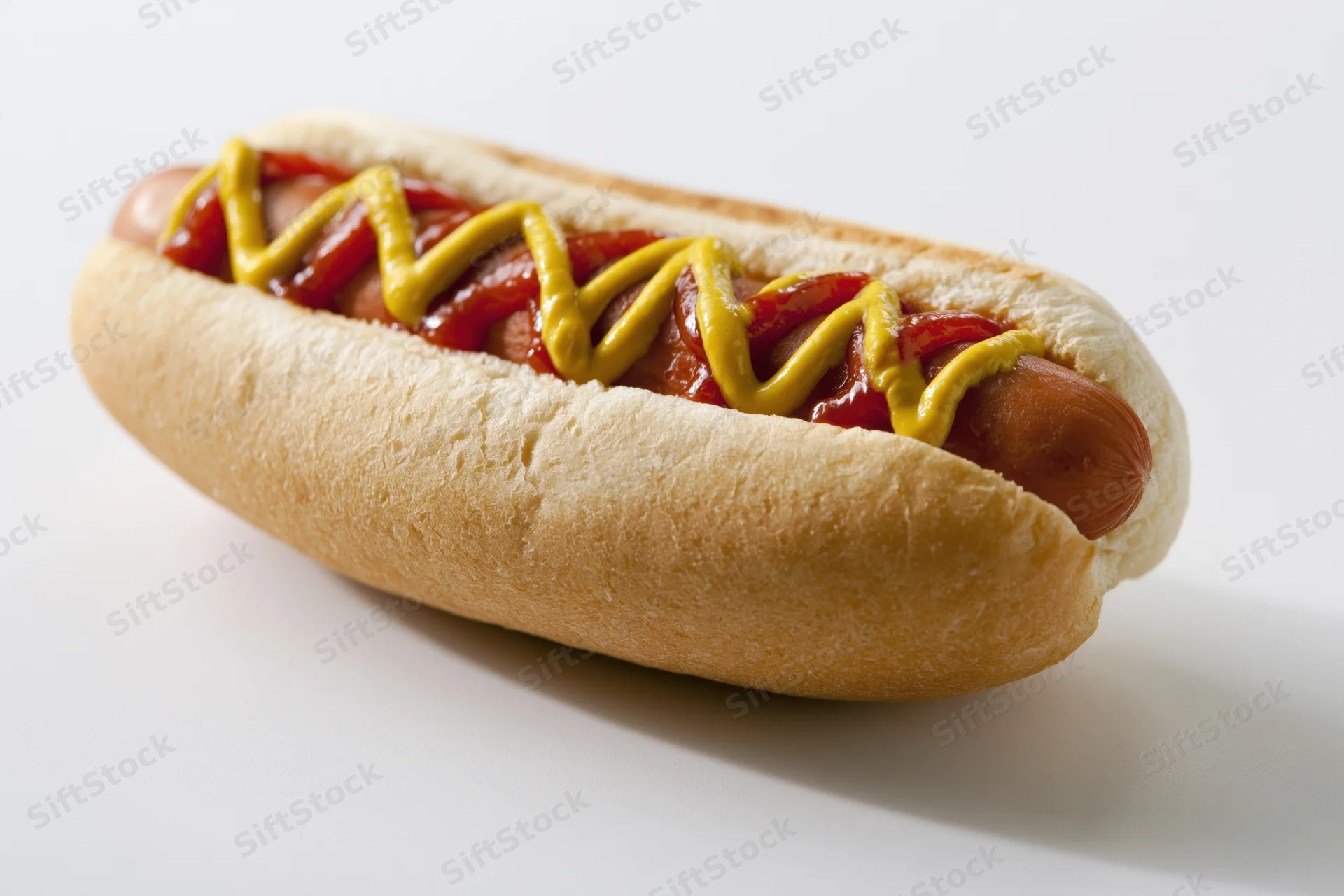 Hot dog in a soft white bun topped with zigzag mustard and ketchup lines on a white surface