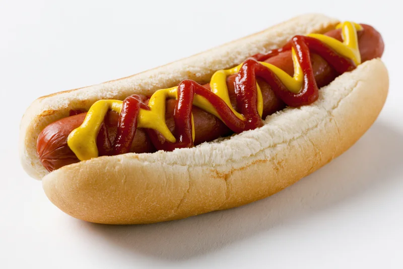 Hot dog in a soft white bun topped with zigzag ketchup and mustard lines on a white surface