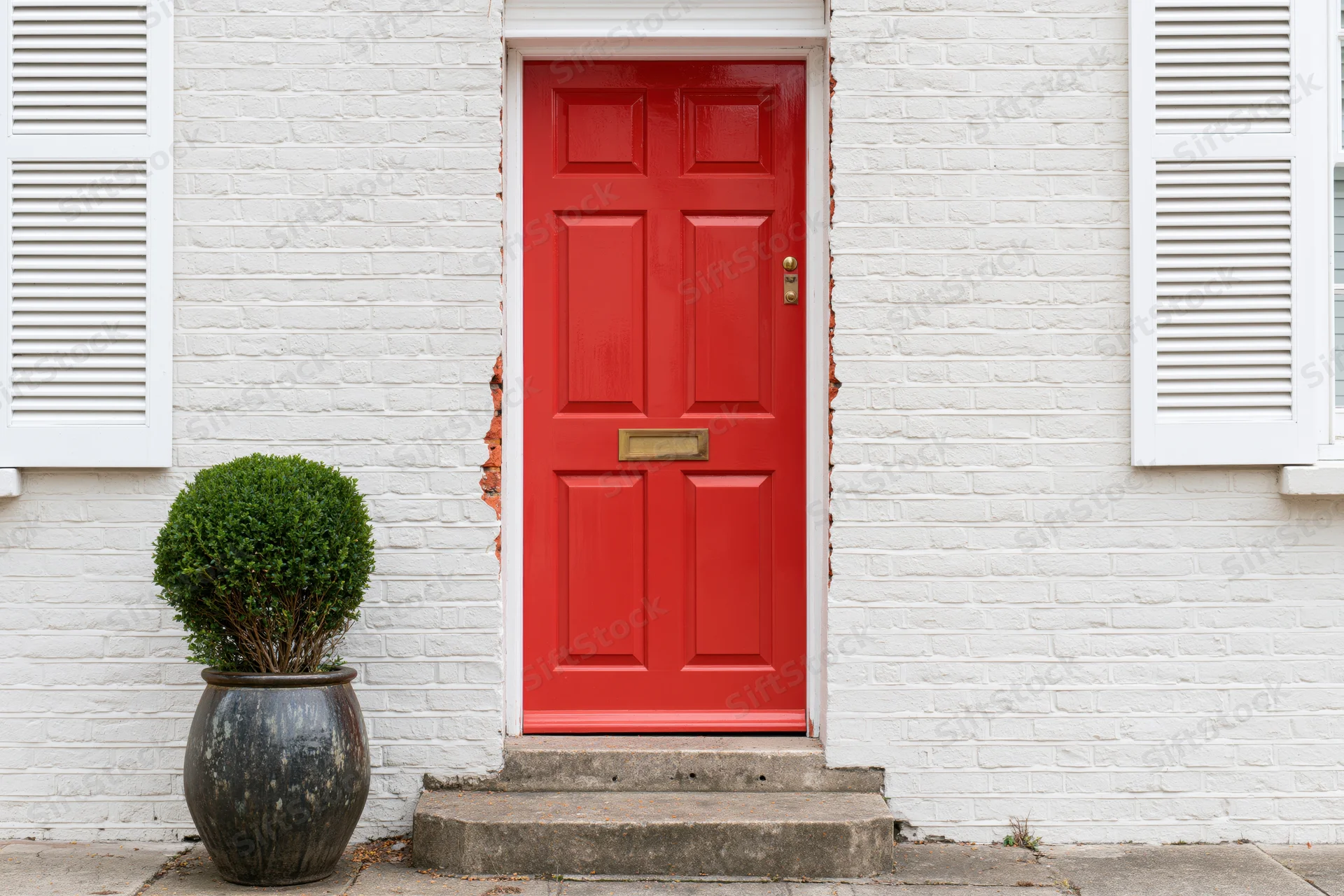 Red six-panel front door with brass hardware on white brick house exterior, flanked by shutters and a potted shrub on concrete steps