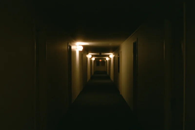 Long empty institutional hallway stretching into darkness with dim wall lights and doors on either side