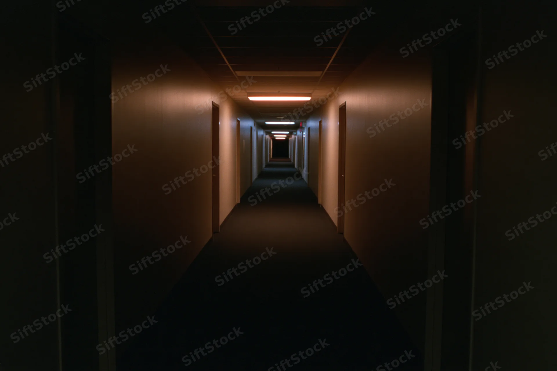 Long empty institutional hallway stretching into darkness with fluorescent lights and doors on both sides