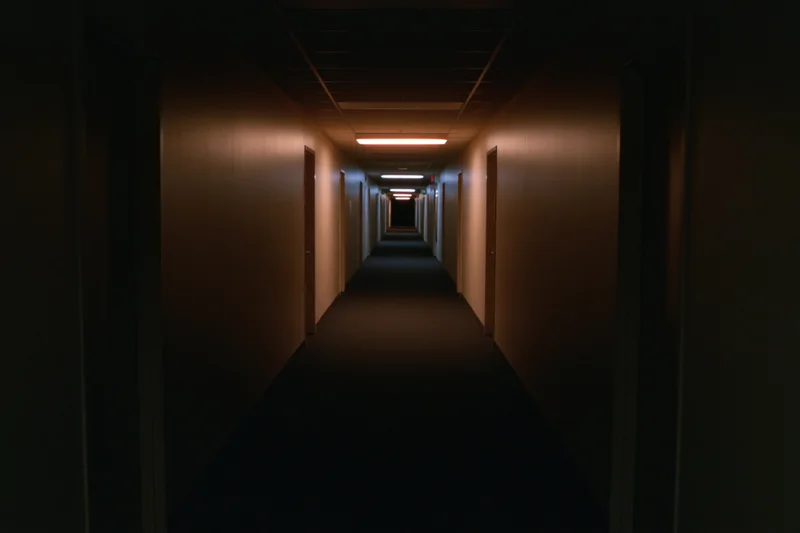 Long empty institutional hallway stretching into darkness with fluorescent lights and doors on both sides