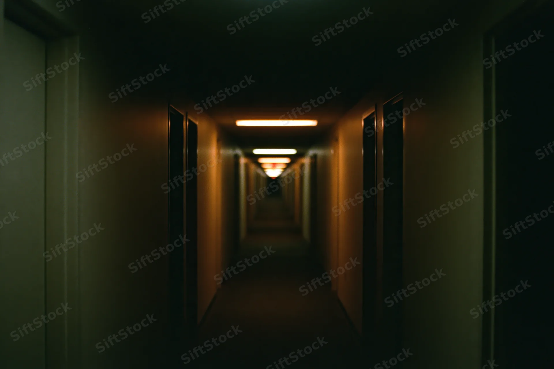 Long empty institutional hallway stretching into darkness with dim fluorescent lighting and repeating doors on both sides