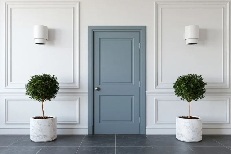 Centered slate-blue interior door flanked by white paneled walls and potted topiaries on gray tiled floor