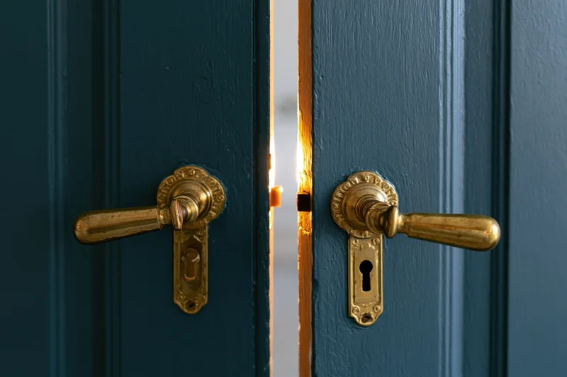 Ornate brass handles on a dark teal double door slightly ajar with light glowing through the gap