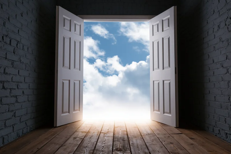 Open white double doors revealing a bright sky and clouds from a dark brick-walled room with wooden floor