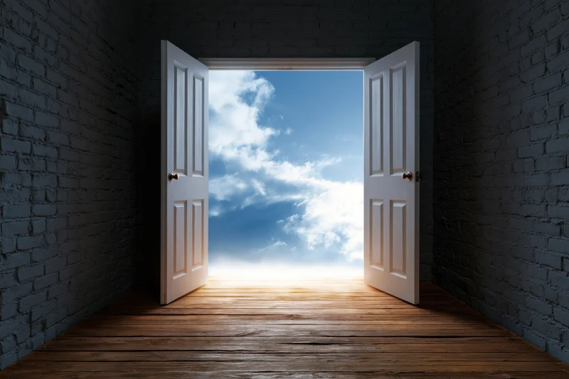 Open white double doors revealing a bright blue sky with clouds beyond a dark brick room