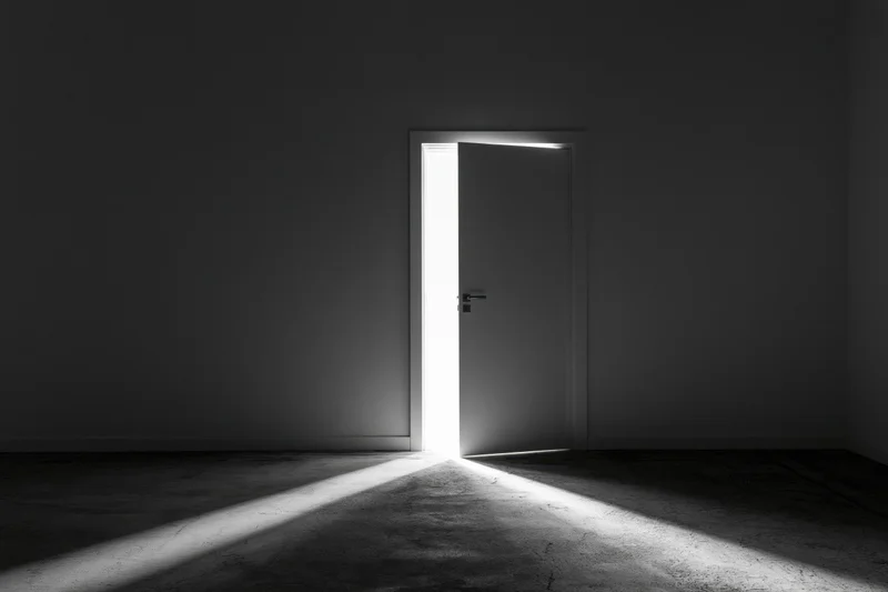 Slightly ajar door in dark empty room letting in bright light beam