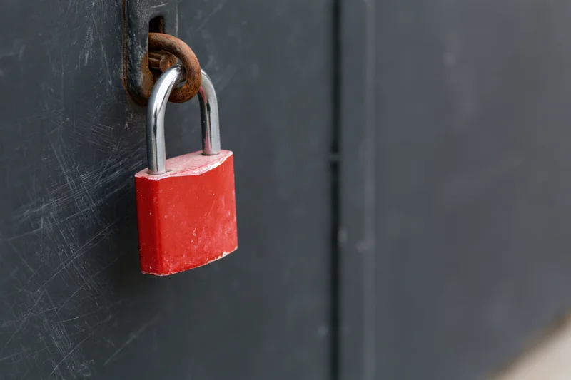Red padlock secured on a rusted metal latch attached to a dark gray door