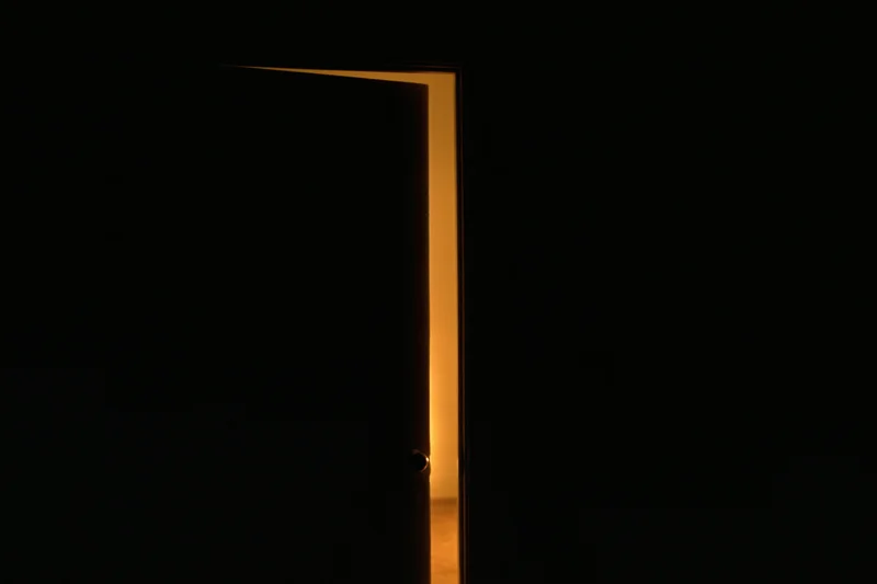 Slightly ajar interior door silhouetted against darkness with warm light glowing from the opening
