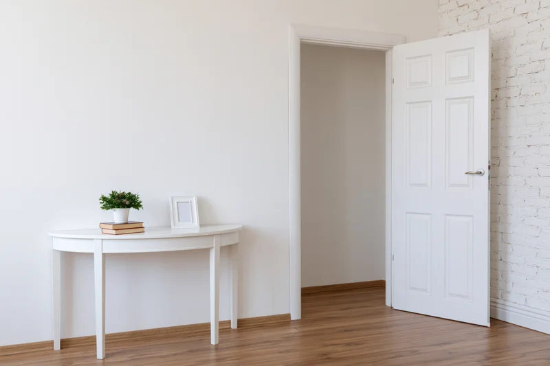White console table with potted plant and picture frame in minimalist entryway with open door and wooden floor