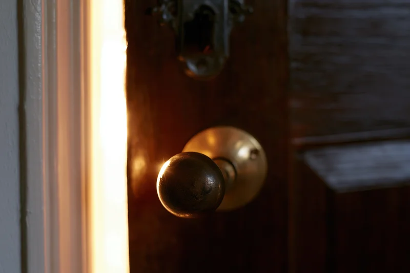 Old dark wooden door slightly ajar with worn brass knob and light glowing through the gap
