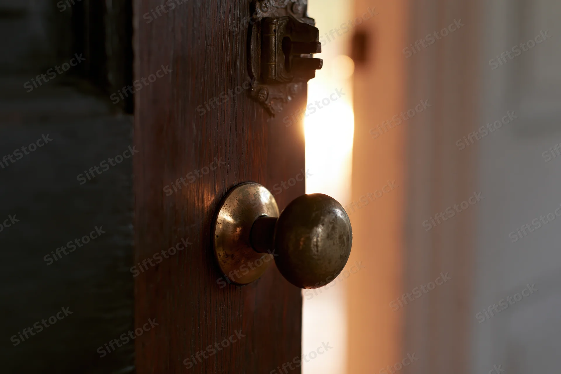 Old dark wooden door slightly ajar with worn brass knob revealing warm light beyond
