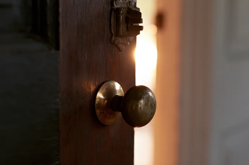 Old dark wooden door slightly ajar with worn brass knob revealing warm light beyond