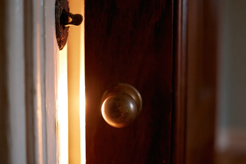 Old dark wooden door slightly ajar with brass knob and light glowing through the crack