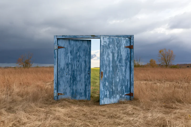 Open weathered blue wooden double door in a dry grass field under cloudy sky