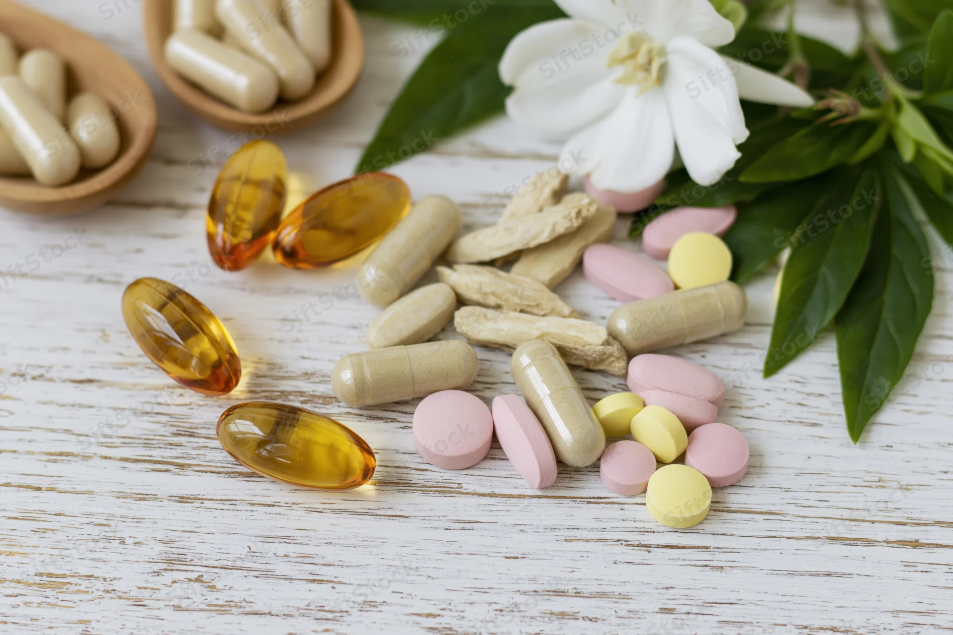 Assorted dietary supplements and vitamins scattered on a white wooden surface with green leaves and a white flower