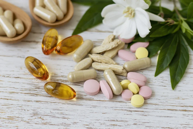 Assorted dietary supplements and vitamins scattered on a white wooden surface with green leaves and a white flower