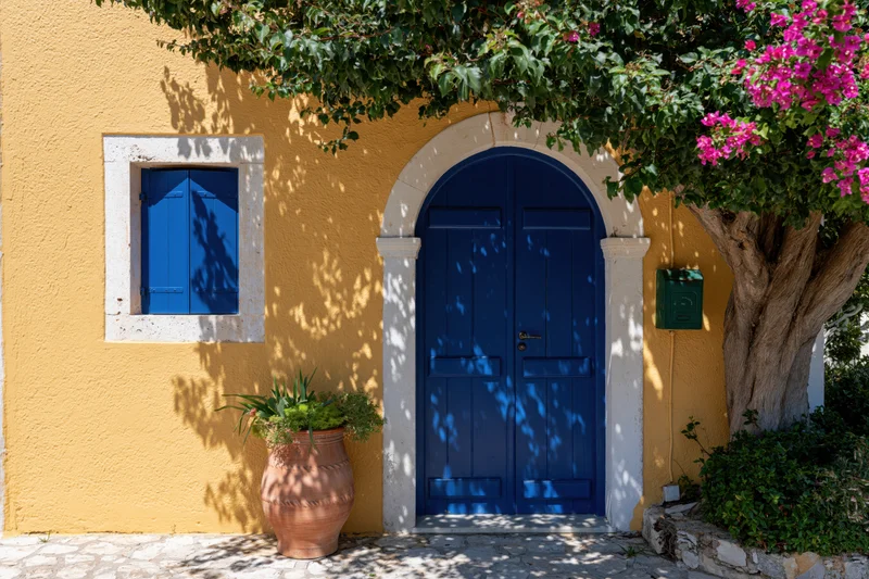 Cobalt blue arched wooden door on a yellow stucco house facade with bougainvillea tree and terracotta pot beside it