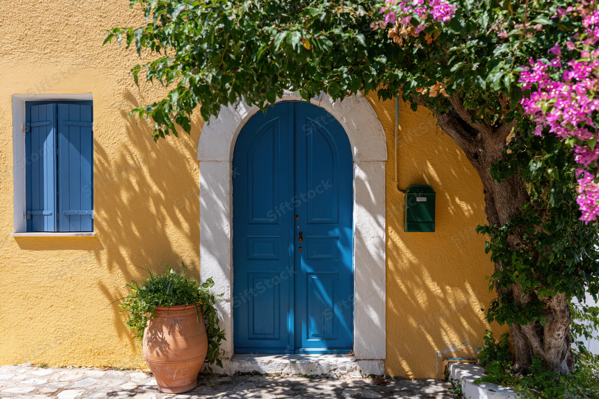 Cobalt blue arched wooden door on a yellow stucco house facade with pink bougainvillea and terracotta planter