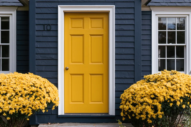 Bright yellow front door with raised panels on a navy blue house flanked by yellow chrysanthemum bushes