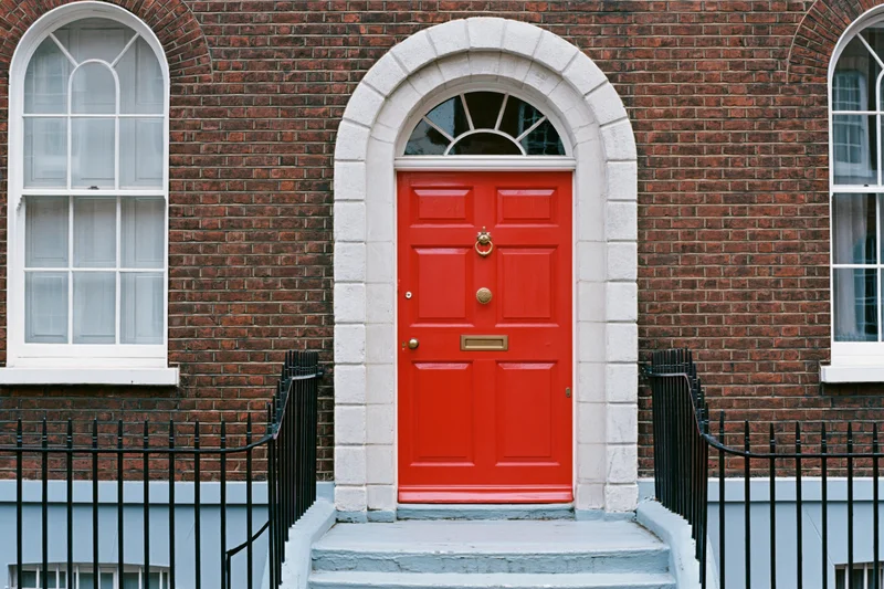 Bright red georgian front door with brass knocker and letterbox set in brick facade with white stone arch and black iron railing