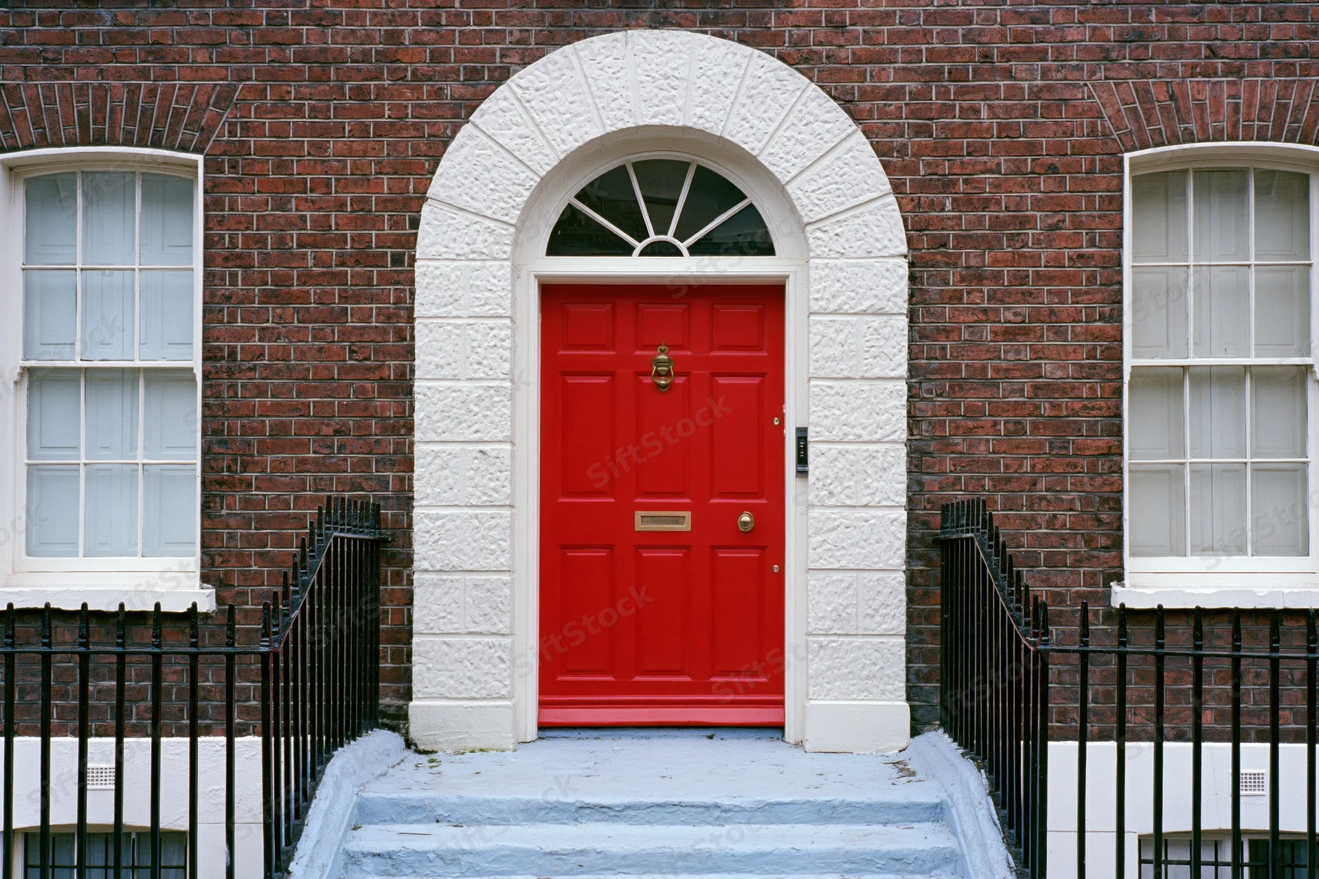 Bright red georgian front door with brass knocker and mail slot set in brick facade with white stone arch and black iron railing