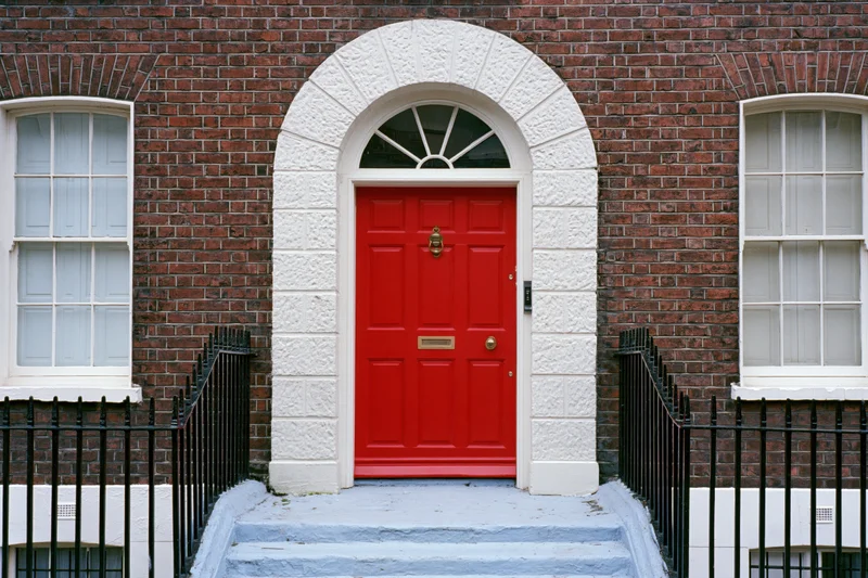 Bright red georgian front door with brass knocker and mail slot set in brick facade with white stone arch and black iron railing