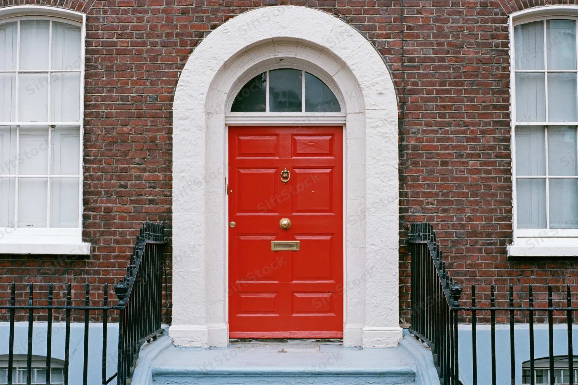 Bright red georgian front door with brass knocker and mailslot set in brick facade with white arched frame and black iron railing
