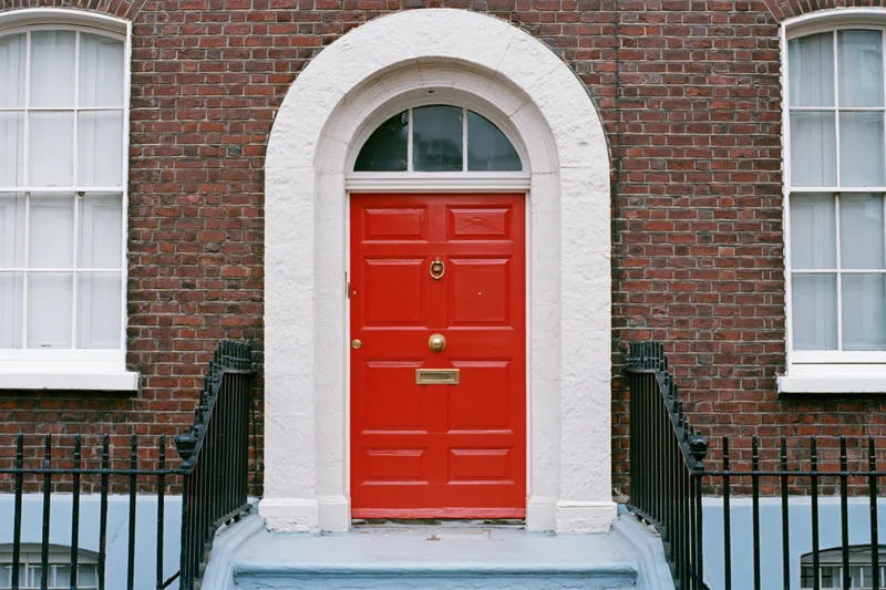 Bright red georgian front door with brass knocker and mailslot set in brick facade with white arched frame and black iron railing