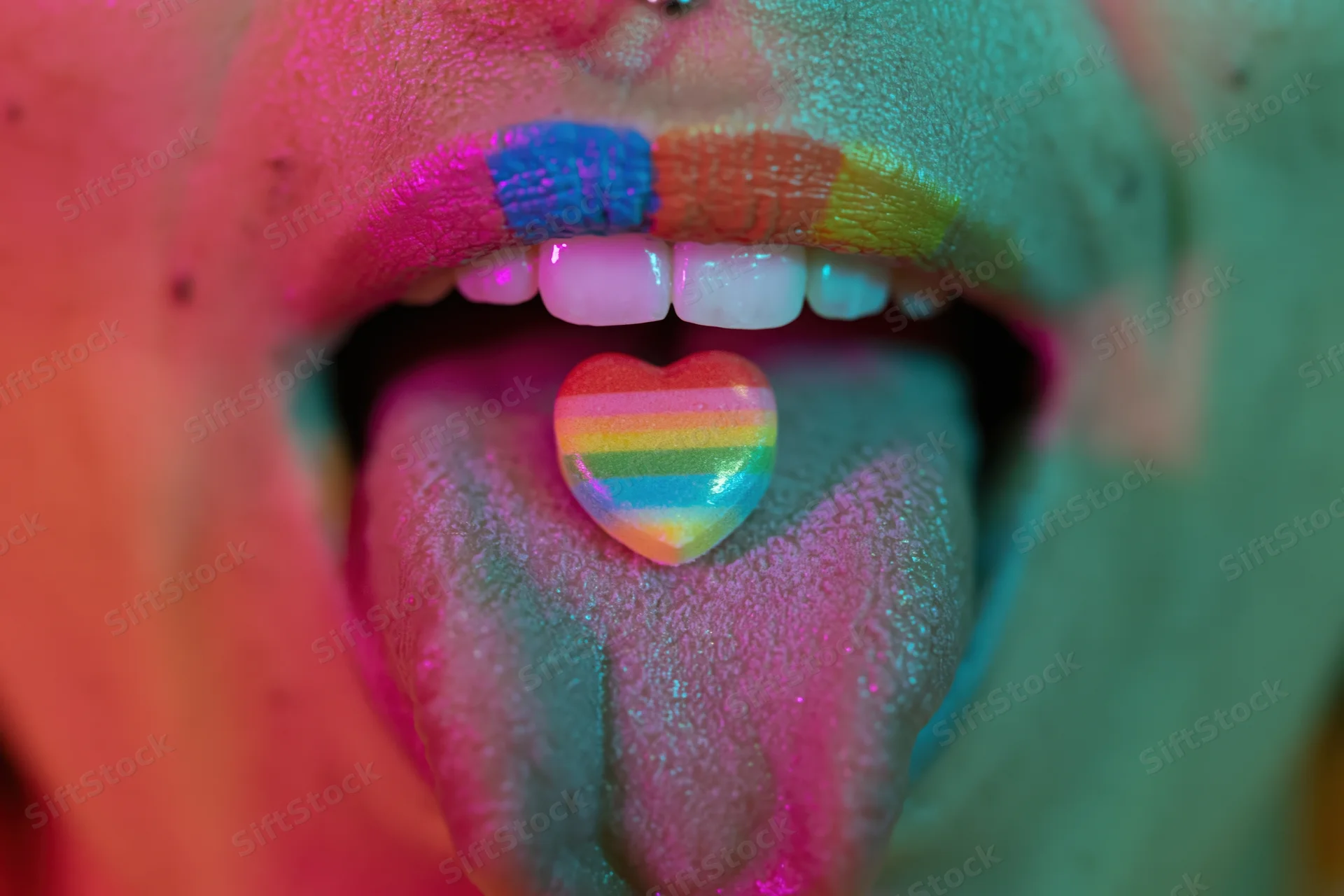 Woman with open mouth showing tongue holding rainbow heart candy, face painted in neon colors